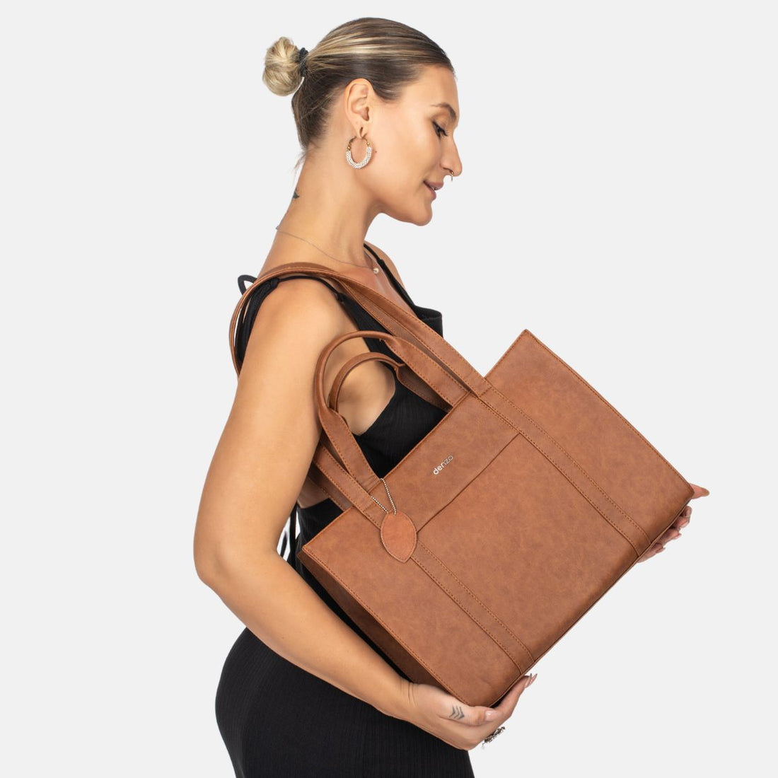 A woman with blond hair tied in a bun is wearing a black sleeveless outfit and holding an elegant everyday tote made of vegan leather over her shoulder. The Lady Leader Tan Tote features an exterior pocket and a small tag, as she gazes downwards against the plain white background.